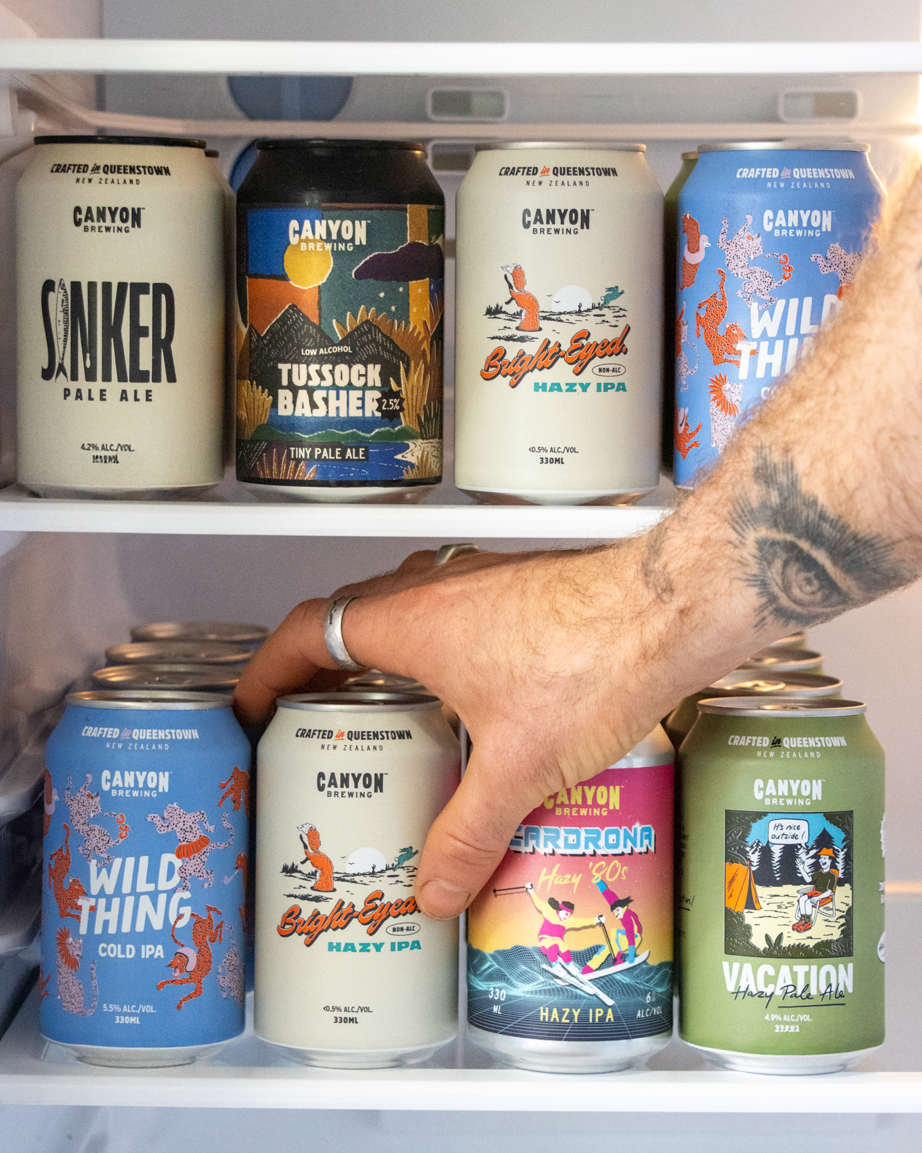 A hand reaches for a can of Canyon Brewing Bright-Eyed on a refrigerator shelf, with other Canyon beer cans displayed on the shelves above.